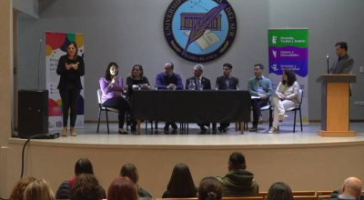 Panel de oradores en el acto de la Universidad Nacional del Sur. Una int&eacute;rprete de Lengua de Se&ntilde;as Argentina (LSA) se encuentra de pie a la izquierda. De fondo, banners institucionales y el escudo de la UNS.